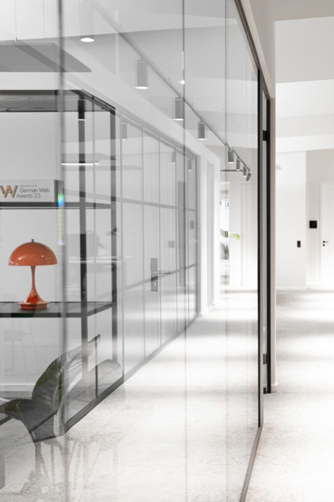 Moderner Büroflur mit Glaswänden, schwarzem Metallrahmen, heller Beleuchtung und oranger Designerlampe im Arbeitsbereich.