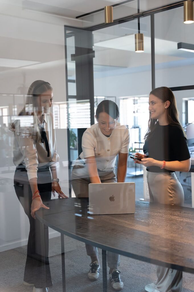 Drei Mitarbeiterinnen der Influencer Marketing Agentur BUZZINTALENTS stehen in einem modernen Besprechungsraum vor einem Laptop.